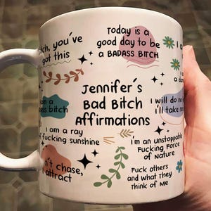 May include: White ceramic mug with the text "Jennifer's Bad Bitch Affirmations" in black. The mug features various affirmations and decorative elements in a handwritten style, including floral and star motifs.