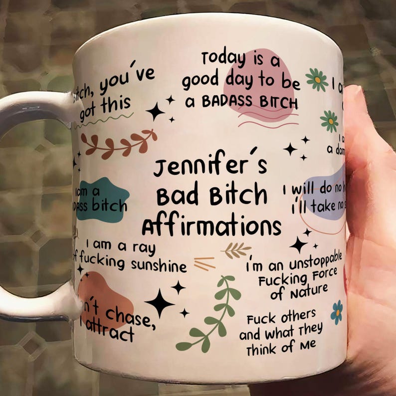 May include: White ceramic mug with the text "Jennifer's Bad Bitch Affirmations" in black. The mug features various affirmations and decorative elements in a handwritten style, including floral and star motifs.