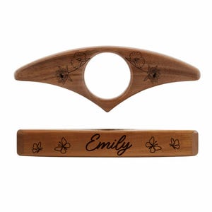 May include: A wooden book holder with a circular opening and a rectangular base. The top piece has floral engravings and the base is engraved with the name "Emily" and butterfly designs. The wood has a natural, warm tone.