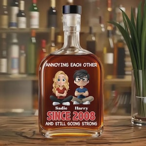 Puede incluir: Una botella de licor de vidrio transparente con un tapón negro, llena de líquido marrón. La etiqueta presenta figuras de dibujos animados y el texto "ANNOYING EACH OTHER SINCE 2008 AND STILL GOING STRONG".