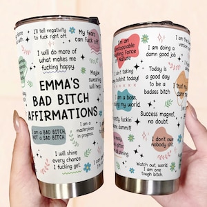 May include: A white stainless steel tumbler with a black lid, featuring the text "EMMA'S BAD BITCH AFFIRMATIONS" and various affirmations in a playful font. The tumbler is decorated with floral and star graphics.