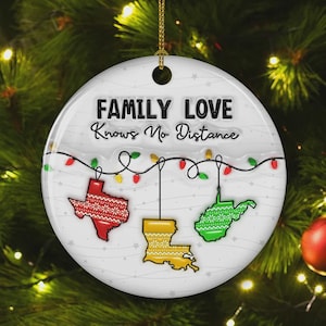 May include: White ceramic Christmas ornament with the text "FAMILY LOVE Knows No Distance." The ornament features red, yellow, and green state outlines with festive patterns, connected by a string of lights. A gold string is attached for hanging.