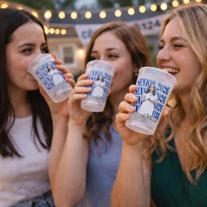 May include: Three clear plastic cups with blue text and a photo of a person are held up by three people. The cups say "SENIOR 2026" and have the name "Patricia" printed on them. The background is blurred with string lights.