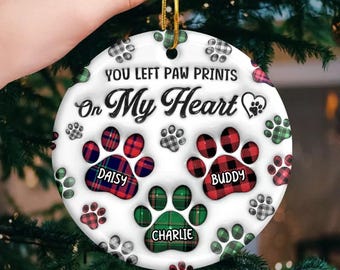 May include: A circular Christmas ornament with the words "YOU LEFT PAW PRINTS On My Heart." It features three paw prints with the names "DAISY," "BUDDY," and "CHARLIE." The paw prints have red, green, and grey plaid patterns. A gold string is attached.