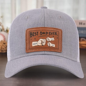 Peut inclure: Casquette de camionneur grise et blanche avec un patch en cuir marron qui dit "Best Dad Ever" avec trois poings serrés et les noms "Daddy", "Emma" et "Andy" sur le patch.