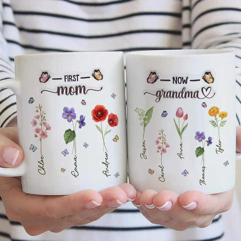 Op de afbeelding: Twee witte keramische mokken met bloemmotieven en de woorden "First Mom" en "Now Grandma". Elke mok heeft namen en illustraties van vlinders en bloemen. De mokken worden door een persoon vastgehouden.