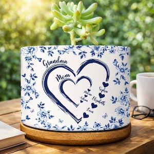 Può includere: Vaso di ceramica bianco con motivo floreale blu e le parole "Nonna" e "Mamma" all'interno di un cuore. I nomi dei membri della famiglia sono elencati sotto. Il vaso è su una base di legno e contiene una pianta succulenta.