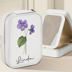 May include: White travel jewelry case featuring a purple flower design and the name "Linda". The case is open, revealing a mirror. The case is made of a smooth material and has a zip closure.