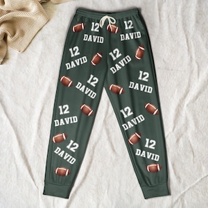 May include: Dark green lounge pants featuring a repeating pattern of brown American footballs and the white text 12 DAVID. The pants have an elastic waistband with a white drawstring and cuffed ankles, showcasing a consistent athletic-themed design.