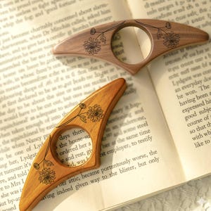 May include: Two wooden thumb page holders with floral designs are displayed on an open book. The page holders are light brown with a circular opening and a curved shape. The floral design is etched in black.