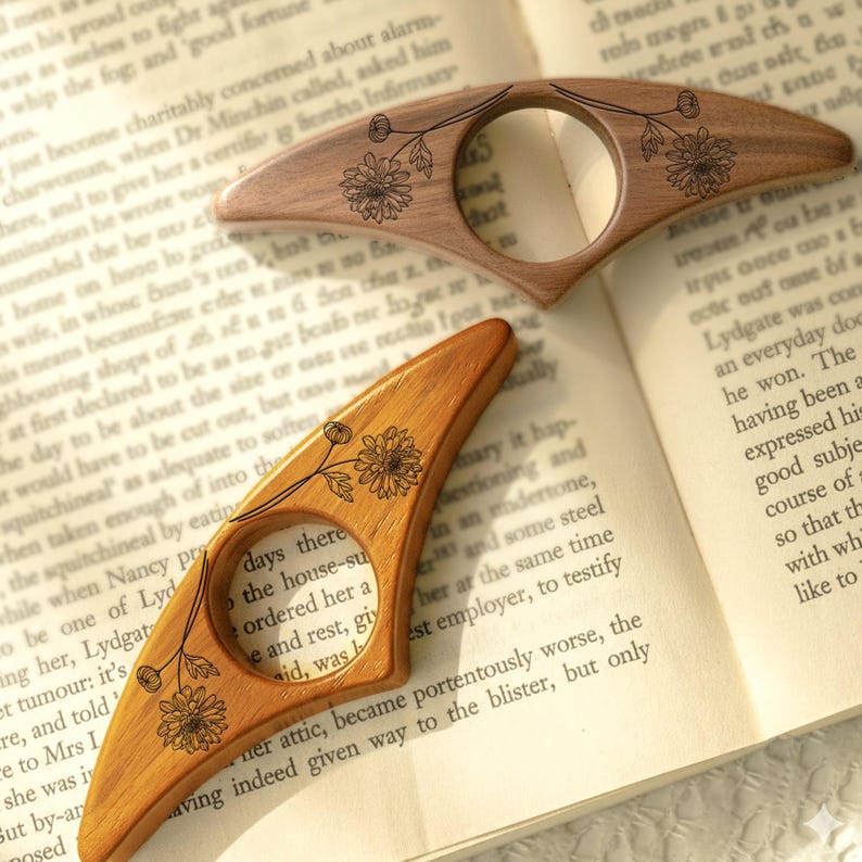 May include: Two wooden thumb page holders with floral designs are displayed on an open book. The page holders are light brown with a circular opening and a curved shape. The floral design is etched in black.