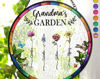 May include: A round stained glass window hanging with a black metal frame. The window has a rainbow border and a white background with the text "Grandma's GARDEN" in black. There are four flowers with names written below them: Caroline, Isabella, Jillian, and Tiffany.