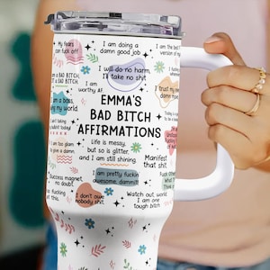 May include: A white insulated tumbler with a handle, featuring the text "EMMA'S BAD BITCH AFFIRMATIONS" and various affirmations. The tumbler has a silver lid and is decorated with floral and star graphics.