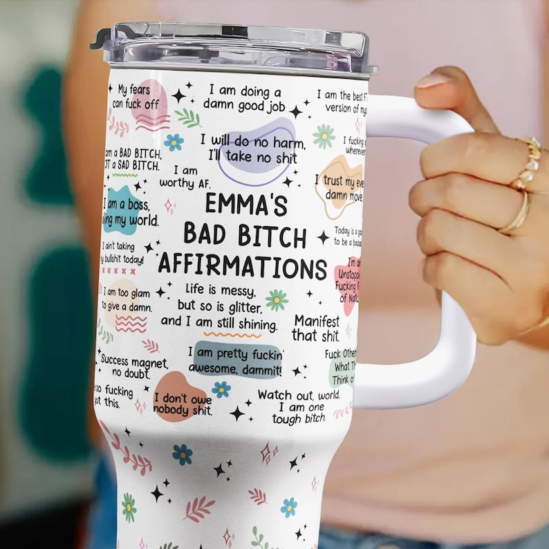 May include: A white insulated tumbler with a handle, featuring the text "EMMA'S BAD BITCH AFFIRMATIONS" and various affirmations. The tumbler has a silver lid and is decorated with floral and star graphics.