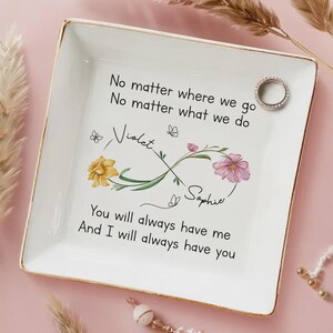 Op de afbeelding: Een witte keramische ringschaal met gouden rand, versierd met bloemen- en vlindermotieven. De schaal draagt de namen "Violet" en "Sophie" en de tekst "No matter where we go..." en "You will always have me..." Een zilveren ring ligt op de schaal.