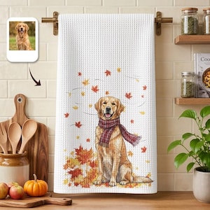 May include: A white waffle-weave kitchen towel featuring a printed illustration of a golden retriever wearing a red and black plaid scarf, surrounded by falling autumn leaves in shades of orange, yellow, and red, with a small photo of the dog above.