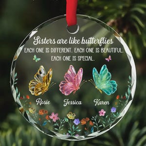 May include: A clear, round glass ornament with a red ribbon. The ornament features the text "Sisters are like butterflies" and the names "Rosie", "Jessica", and "Karen". Three colorful butterflies and floral accents decorate the ornament.