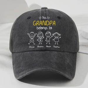 Peut inclure: Casquette de baseball gris foncé avec l'inscription jaune "This Grandpa belongs to" et les noms Oliver, Amelia, Noah et Sophia sous des personnages stylisés. La casquette a une visière incurvée.