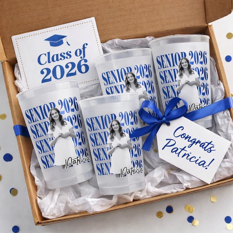 May include: A graduation gift box containing four clear plastic cups printed with "SENIOR 2026" and a photo of a woman. The box also includes a card that says "Class of 2026" and a tag that says "Congrats Patricia!" A blue ribbon is tied around the box.