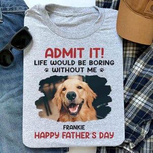 May include: A gray t-shirt with the text "ADMIT IT! LIFE WOULD BE BORING WITHOUT ME" and a photo of a golden retriever. The shirt also says "FRANKIE HAPPY FATHER'S DAY". Sunglasses, a hat, and a plaid shirt are in the background.