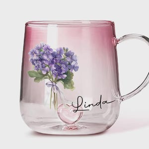 May include: A clear glass mug with a pink ombre effect. The mug features a floral design of purple flowers and the name "Linda" in script. A heart-shaped detail is at the base of the mug.