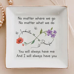Op de afbeelding: Een vierkant, wit keramisch sieradenschaaltje met een gouden rand. De schaal heeft een oneindigheidssymbool met bloemen- en vlinderaccenten, de namen "Isabella" en "Jennifer" en de tekst "No matter where we go..." en "You will always have me..."