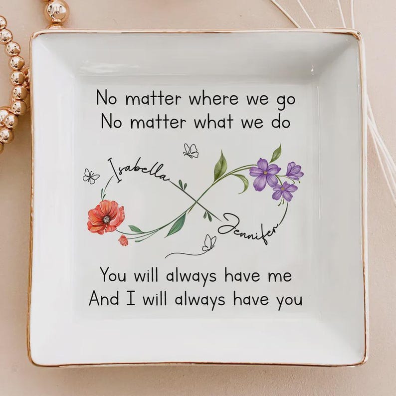 Op de afbeelding: Een vierkant, wit keramisch sieradenschaaltje met een gouden rand. De schaal heeft een oneindigheidssymbool met bloemen- en vlinderaccenten, de namen "Isabella" en "Jennifer" en de tekst "No matter where we go..." en "You will always have me..."