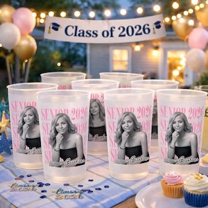 May include: A collection of frosted plastic cups with a photo of a woman, the text "SENIOR 2026" in pink, and the name "Barbara". A banner reads "Class of 2026". The scene includes cupcakes and balloons.