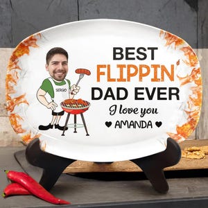 Peut inclure: Un plat de service ovale blanc avec une bordure à motif de flammes. Il présente une illustration de dessin animé d'une personne en tablier blanc faisant griller des aliments, avec le texte "BEST FLIPPIN DAD EVER" et "I love you AMANDA".