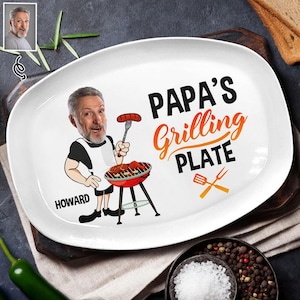 Könnte beinhalten: Weiße ovale Platte mit dem Text "PAPA'S Grilling PLATE" in Schwarz und Orange. Auf der Platte befindet sich eine Cartoon-Illustration eines grillenden Mannes. Der Name "HOWARD" ist auf der Platte aufgedruckt.