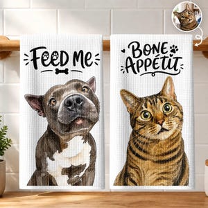 May include: Two white waffle-weave kitchen towels featuring watercolour-style pet illustrations. The left towel shows a dark brown and white dog with the text "Feed Me" and a bone icon. The right towel shows a brown tabby cat with the text "Bone Appétit".