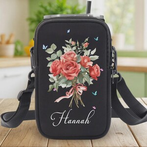May include: Black water bottle carrier with a floral design. The carrier features a bouquet of red roses, green leaves, and small blue butterflies. The name "Hannah" is written in cursive script below the flowers. It has a black adjustable strap.