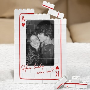 May include: A rectangular photo frame constructed from white interlocking plastic building blocks, designed to resemble an Ace of Hearts playing card. It features a black and white photograph of two people and the red text How lucky are we?