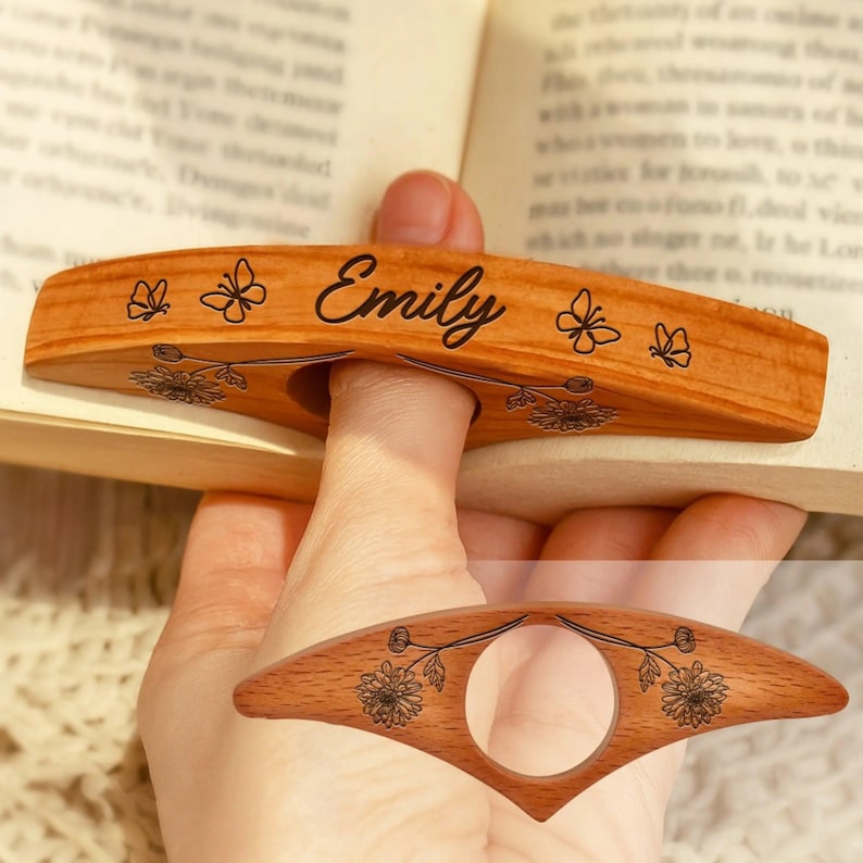 May include: A wooden book page holder with the name "Emily" engraved in black script. The holder features butterfly and floral designs. The holder is designed to fit over a finger, keeping the book open.