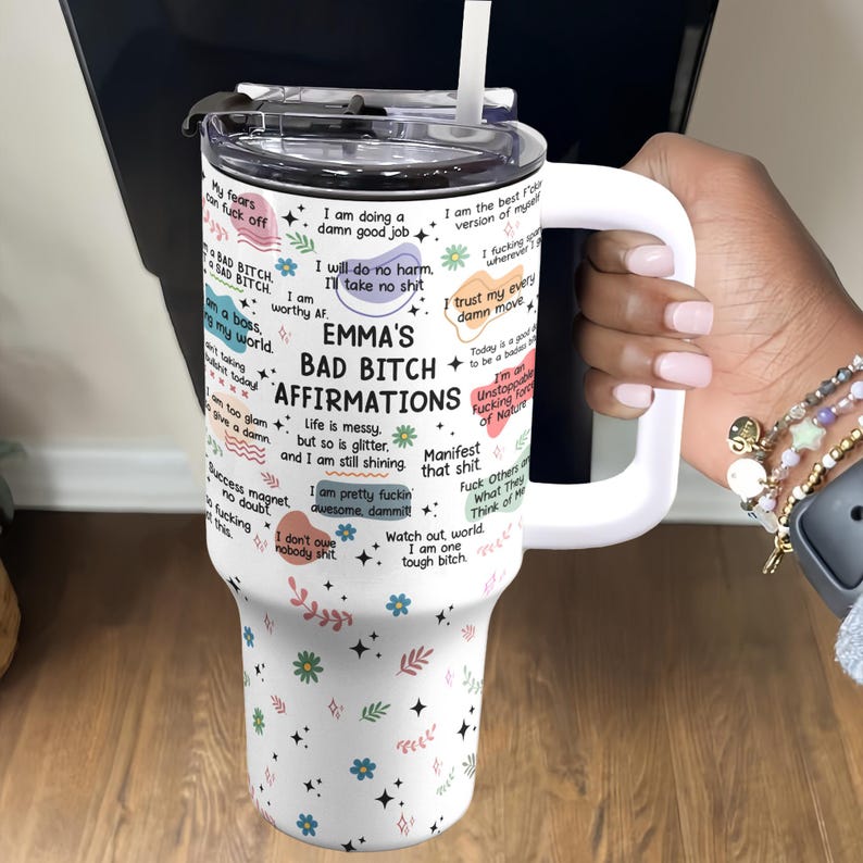 May include: A white tumbler with a handle, featuring the text "EMMA'S BAD BITCH AFFIRMATIONS" and various affirmations. The tumbler has a white lid and a straw. Floral and star patterns decorate the tumbler.
