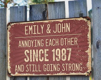 May include: A rectangular, vintage-style metal sign with the text "EMILY & JOHN ANNOYING EACH OTHER SINCE 1987 AND STILL GOING STRONG" in white lettering on a maroon background. The sign is mounted on a weathered wooden fence.