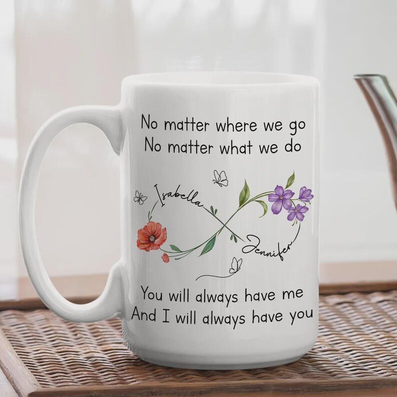 Op de afbeelding: Witte keramische mok met een handvat, met de tekst "No matter where we go, no matter what we do." De mok heeft een oneindigheidssymbool met bloemen- en vlinderaccenten, en de namen "Isabella" en "Jennifer".