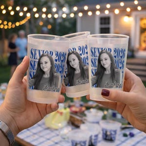 May include: Three clear plastic cups with a photo of a young woman and the text "SENIOR 2026" in blue. The cups are held up in front of a blurred outdoor background with string lights.