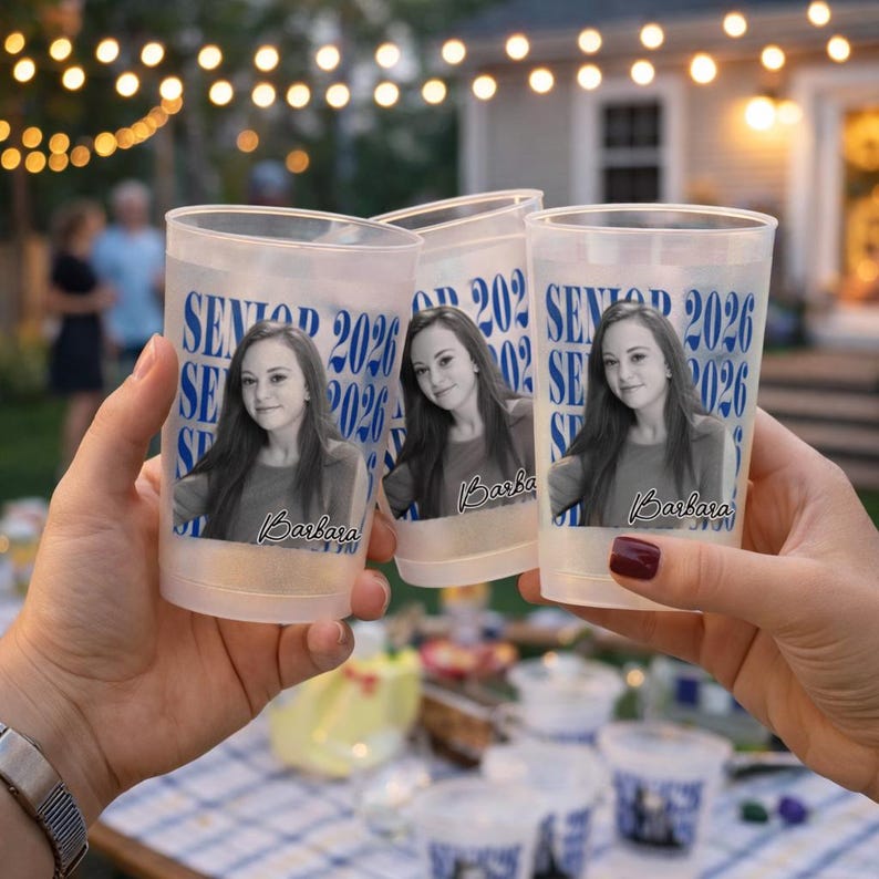 May include: Three clear plastic cups with a photo of a young woman and the text "SENIOR 2026" in blue. The cups are held up in front of a blurred outdoor background with string lights.