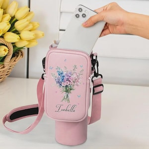 May include: Pink crossbody phone bag with a floral bouquet design and the name "Isabella". The bag has a matching pink strap and a cylindrical bottom section, possibly for a water bottle. A silver smartphone is being placed inside.