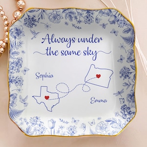 May include: Square white ceramic plate with blue floral and butterfly pattern. The plate has a gold rim and features the text "Always under the same sky" with two states and names, Sophia and Emma, connected by a dotted line.