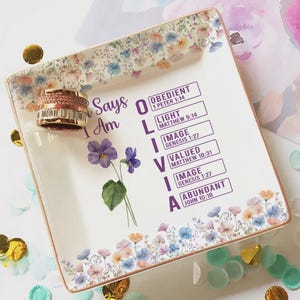 May include: A square, floral-decorated ceramic tray with the name "Olivia" and inspirational words. A gold and crystal ring rests on the tray. The tray is adorned with a floral border and scattered confetti.