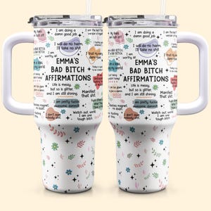 May include: Two white travel mugs with handles, featuring the text "EMMA'S BAD BITCH AFFIRMATIONS" and various affirmations. The mugs have a floral pattern with stars and colorful accents. Each mug has a white lid and a straw.
