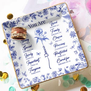 May include: A square, white ceramic trinket tray with a gold rim, featuring blue floral patterns and inspirational text. A rose gold ring sits on the tray. The tray includes the words "You Are" and other positive affirmations.