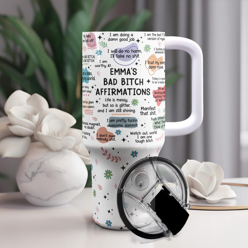 May include: A white tumbler with a handle and a black lid. The tumbler is decorated with affirmations and the text "EMMA'S BAD BITCH AFFIRMATIONS". Floral and leaf designs are scattered around the tumbler.