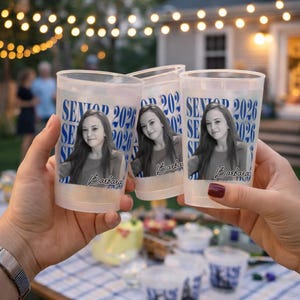 May include: Three clear plastic cups with a photo of a young woman, the name "Barbara", and the text "SENIOR 2026" in blue. The cups are held up in a toast, with a blurred background of a backyard party.