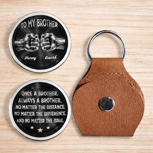 Può includere: Un set di portachiavi personalizzato con due gettoni rotondi in metallo e un portachiavi in pelle marrone. I gettoni hanno sfondi neri con testo e illustrazioni bianche. Uno dice "TO MY BROTHER" con due pugni e nomi.