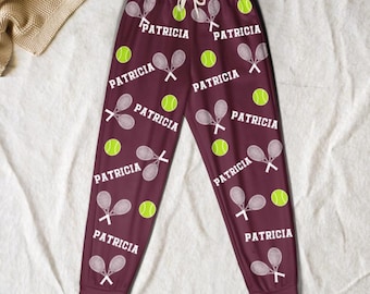 May include: Burgundy-colored sweatpants with a white drawstring, featuring a pattern of tennis rackets, tennis balls, and the name "PATRICIA" in white. The design is repeated across the pants.