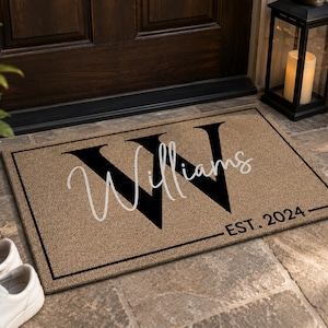 Puede incluir: Un felpudo rectangular de color tostado con un borde negro. La alfombra presenta una letra W grande y negra en el fondo, con el nombre Williams escrito en cursiva blanca sobre ella y el texto EST. 2024 en negro en la esquina inferior derecha.