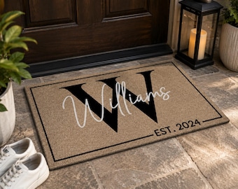 May include: A rectangular tan doormat with a black border. The mat features a large black letter W in the background, with the name Williams written in white cursive script across it and the text EST. 2024 in black in the bottom right corner.